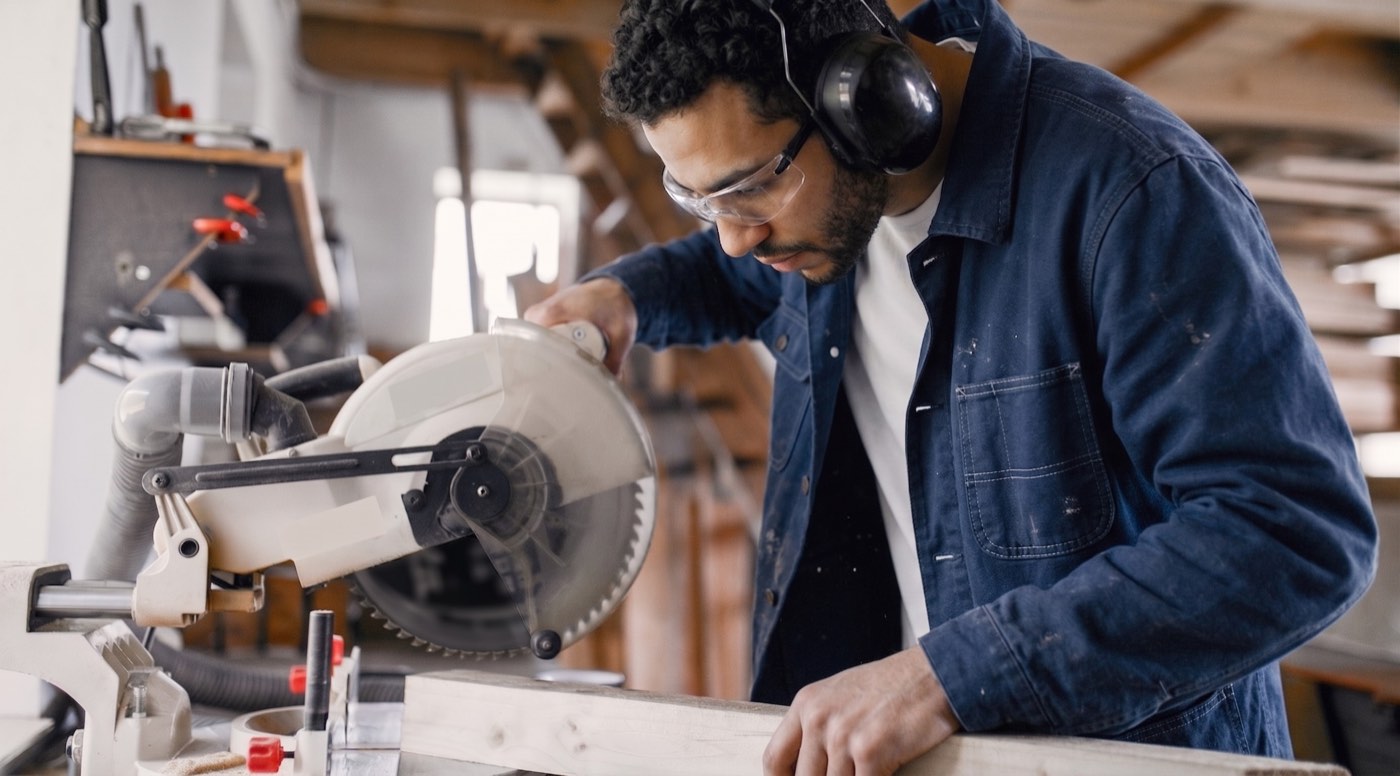 Menuisier équipé de lunettes et d'un casque anti-bruit découpant une planche à la scie à onglet dans son atelier
